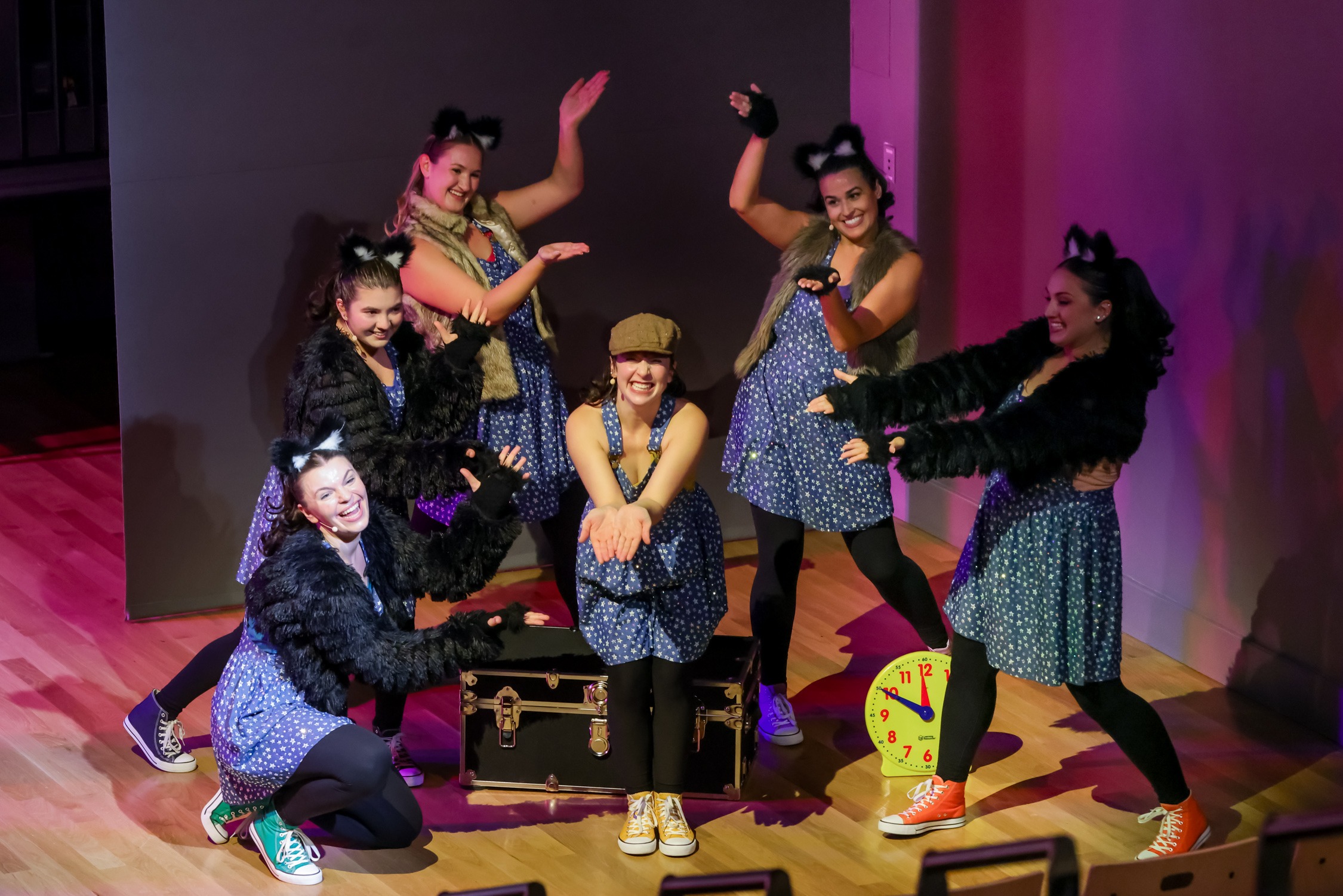 A group of six performers in blue dresses and black furry sleeves pose energetically on stage. A large clock pointed at 11 o'clock and a black trunk are in the background.