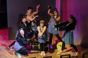 A group of six performers in blue dresses and black furry sleeves pose energetically on stage. A large clock pointed at 11 o'clock and a black trunk are in the background.