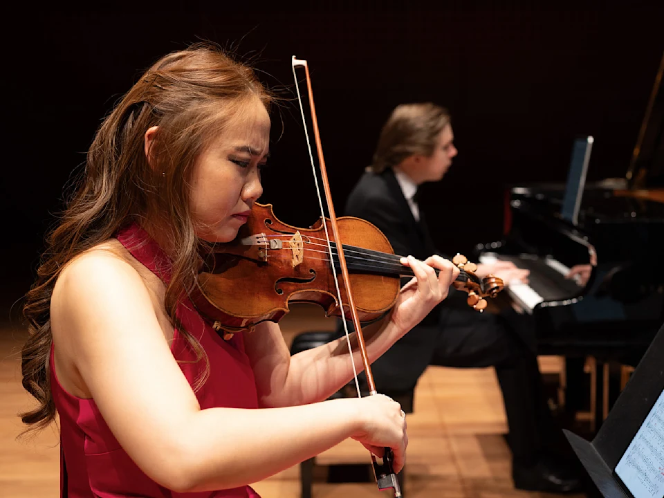 A woman plays violin on stage while a man plays piano in the background. Both are focused on their performances.