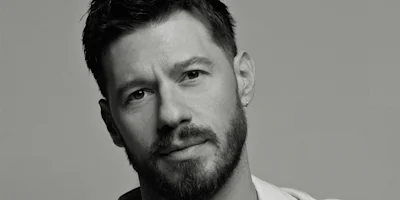 Black and white portrait of a man with short dark hair, a beard, and an earring, looking directly at the camera with a neutral expression.