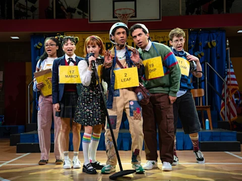 Six young contestants stand on a stage with numbered placards around their necks, gathered around a microphone at a spelling bee event.