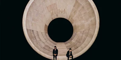 Two people sit on chairs in front of a large, circular wooden structure with a hollow center, against a black background.