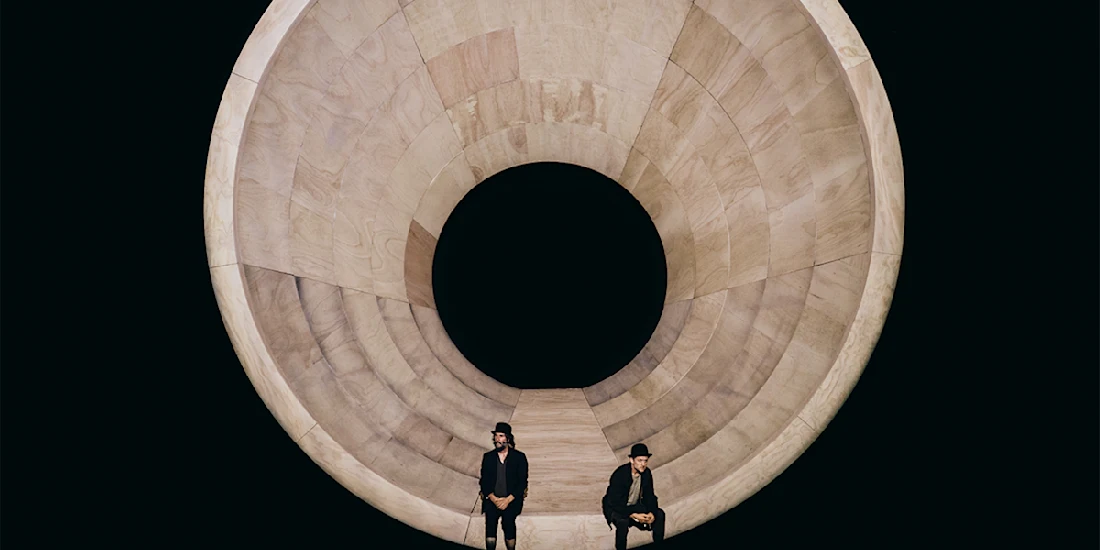 Two people sit on chairs in front of a large, circular wooden structure with a hollow center, against a black background.