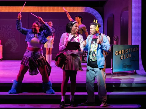 Four young actors perform on stage in colorful costumes; two talk at center while two others dance energetically behind them near a "Christian Club" sign.