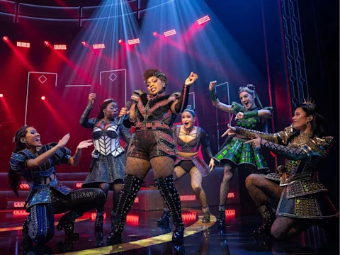 Production photo of SIX in Costa Mesa showing six performers in elaborate, colorful costumes sing and dance energetically on a stage with dramatic lighting and a vibrant set.