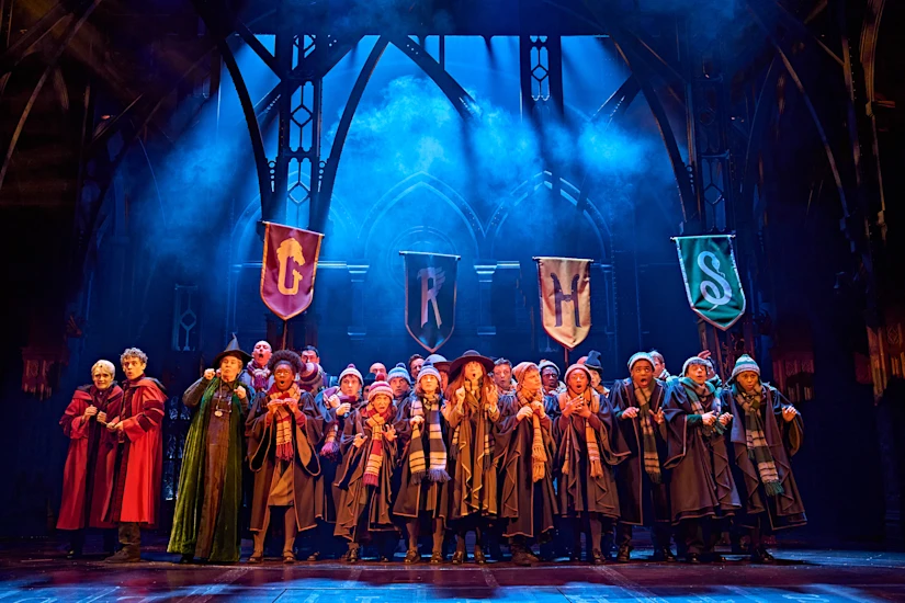A large cast in school robes and scarves performs on a theater stage beneath four house banners.
