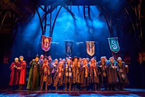 A large cast in school robes and scarves performs on a theater stage beneath four house banners.