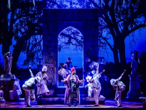 A performer sings on stage in front of an ornate archway, flanked by dancers in white costumes and statues; a backdrop depicting trees and a night sky enhances the theatrical setting.