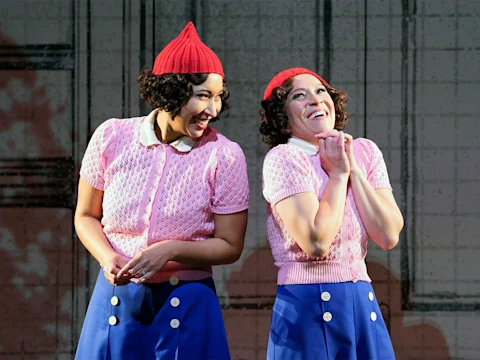 Two people wear matching red hats, pink short-sleeve sweaters, white collars, and blue skirts on stage. One smiles widely while the other clasps their hands under the chin.