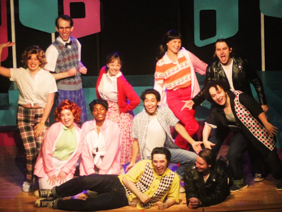 Production photo of Grease in Illinois featuring the cast in colorful, retro costumes pose cheerfully on a stage, some standing and some kneeling or sitting, with vibrant set pieces in the background.