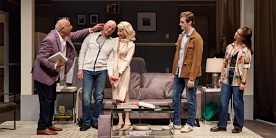 Five people stand in a modern living room set on stage; three are grouped together at center while two others stand apart, observing the scene.