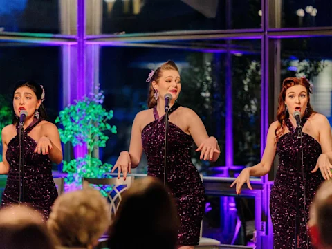 Three women in matching sequined dresses sing into microphones on stage, gesturing expressively with their hands.