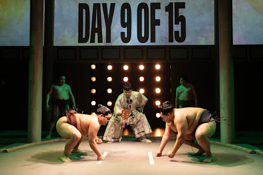 Two sumo wrestlers face off in front of a referee and an audience. A sign above reads "Day 9 of 15.