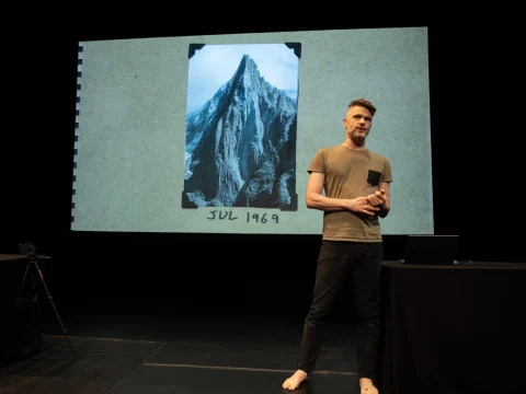 A person stands on stage giving a presentation with a photograph of a mountain labeled "July 1969" displayed on a screen behind them.