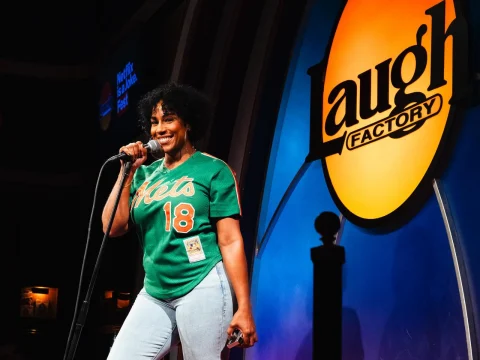 A person in a green "Mets" jersey stands on stage at the Laugh Factory comedy club, holding a microphone and smiling.