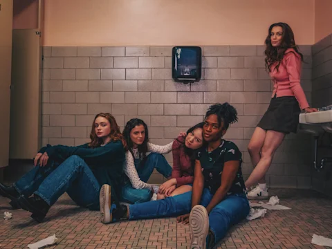 Five people are sitting and standing in a tiled bathroom. They are dressed casually. Some paper towels are scattered on the floor.