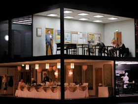 A two-story building with the upper floor showing a classroom and the lower floor set up for a formal dinner event; people are present in both scenes.