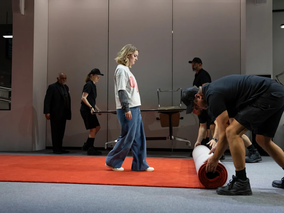 A woman in casual clothes stands on a red carpet as crew members unroll it and move furniture in a modern indoor setting.