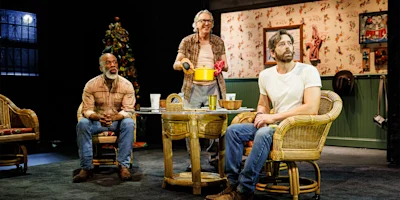 Three men on a stage. One sits in a rattan chair, looking left. Another stands, holding a yellow pot. The third sits, looking forward.
