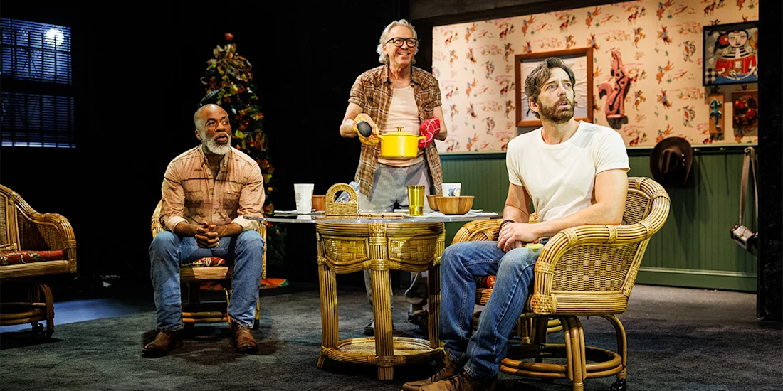 Three men on a stage. One sits in a rattan chair, looking left. Another stands, holding a yellow pot. The third sits, looking forward.