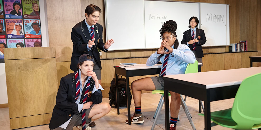 Four actors playing students in school uniforms are in a classroom setting; one sits at a desk appearing thoughtful while the others stand around, with one writing on the whiteboard in the background.