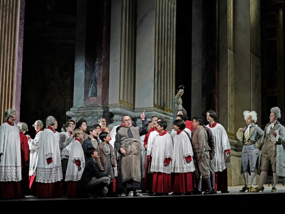 Production photo of Tosca, showing a group of performers in traditional costumes are on stage, with several dressed as choir boys, gathered around a central figure in a stone building setting.