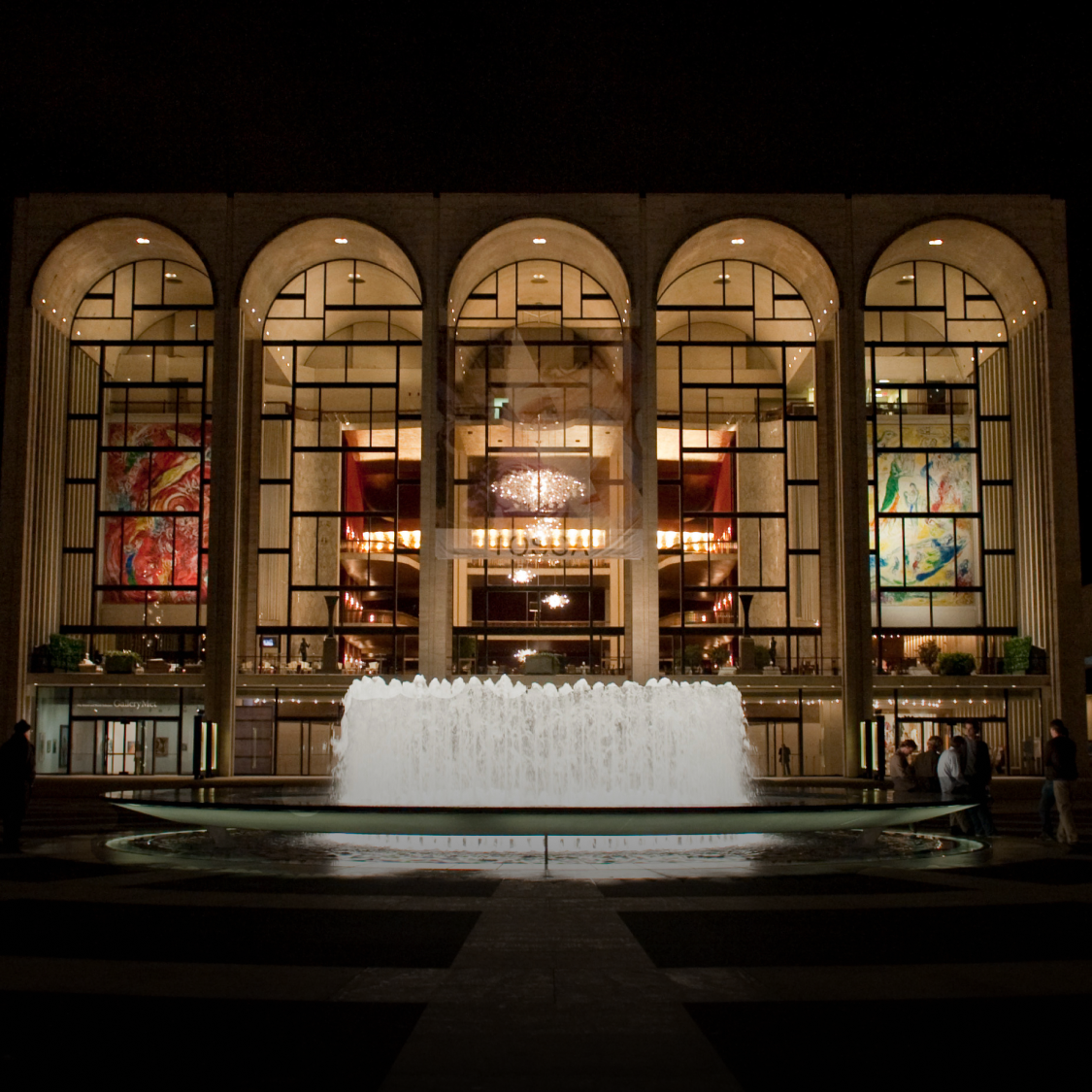 Night view of a grand building with large arched windows, colorful banners, and a central illuminated fountain in front.