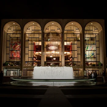 Night view of a grand building with large arched windows, colorful banners, and a central illuminated fountain in front.