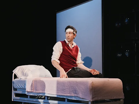 Production photo of Gruesome Playground Injuries in New York, showing a man wearing a head bandage and red vest sits on a hospital bed with a pink blanket, looking to the side against a dark background.