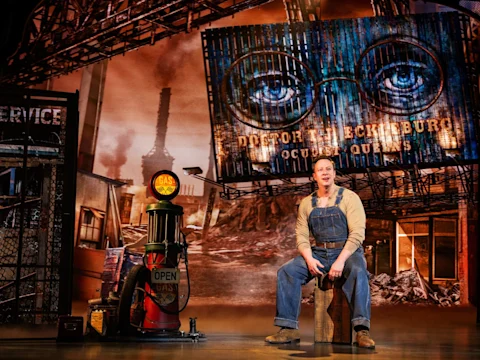 A man in overalls sits on a wooden crate onstage, with a gas pump and a large billboard featuring glasses and eyes behind him, in a theatrical set resembling an industrial area.