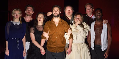 Eight people in period costumes stand together, looking forward with serious and concerned expressions under dramatic stage lighting.