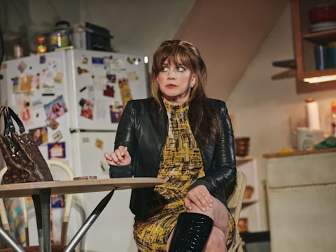 A woman in a yellow patterned dress and black jacket sits at a kitchen table, with a refrigerator covered in magnets and papers in the background.