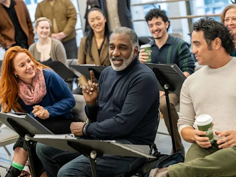 A group of people sit in chairs with scripts on music stands, smiling and engaging in a discussion; one person gestures while speaking.