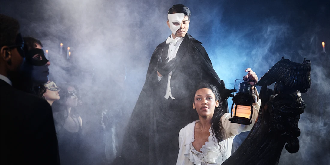 A man in a black cape and half mask stands behind a woman in white holding a lantern, surrounded by people in masquerade costumes and mist.