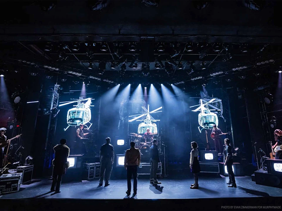 Production photo of Rolling Thunder in New York showing a stage performance with actors standing in a line, dramatic blue lighting, and large digital images of helicopters displayed in the background.