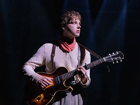 A person wearing a light-colored shirt and red neckerchief stands under stage lights, playing an electric guitar.