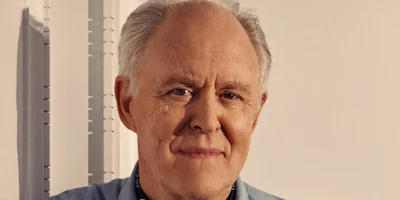 An older man with short gray hair and a light blue shirt stands indoors against a neutral background, looking at the camera.