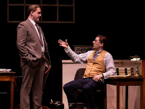 Two men in vintage office attire converse; one stands with hands in pockets while the other, seated at a desk, gestures openly. A lamp, books, and window frame are visible in the background.