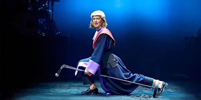 A performer in a judge’s costume kneels on stage, holding a microphone stand horizontally, under blue stage lighting.