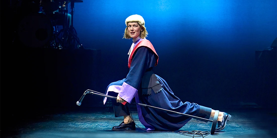 A performer in a judge’s costume kneels on stage, holding a microphone stand horizontally, under blue stage lighting.