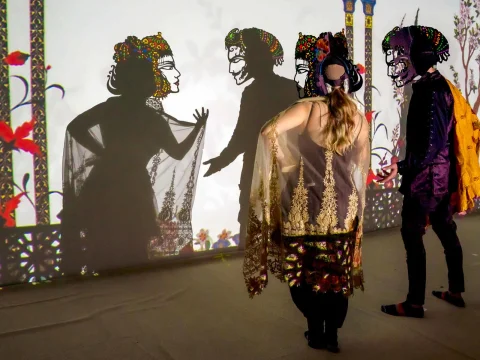 Three people in colorful costumes stand in front of a screen, casting elaborate shadows during a shadow puppet performance.