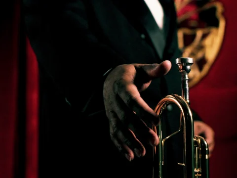 A musician in a black suit reaches for a brass trumpet with a red and gold background.