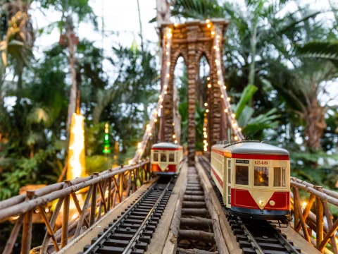 Model trains on parallel tracks cross a miniature bridge adorned with string lights, surrounded by lush plants.