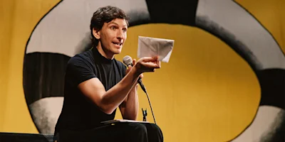 A person in a black shirt holds a microphone and a folded paper, speaking on stage in front of a large yellow and black circular backdrop.