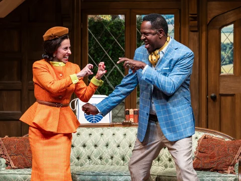 Two actors on stage in an energetic pose, one wearing an orange outfit and the other in a light blue suit. They appear engaged in a lively theatrical scene in a living room setting.