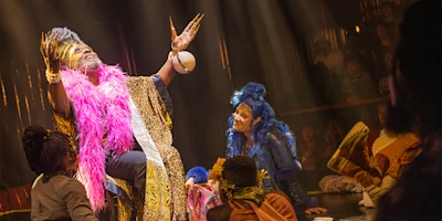 A performer in vibrant, animal-print clothing and a pink feather boa gestures dramatically onstage, surrounded by others in colorful costumes and wigs, under theatrical lighting.