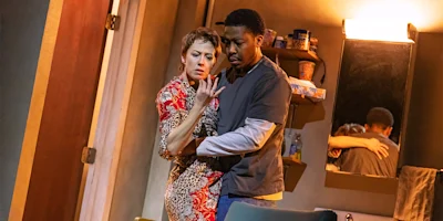 A woman in a patterned dress and a man in scrubs stand closely in a dimly lit room, with their reflection visible in the mirror behind them.