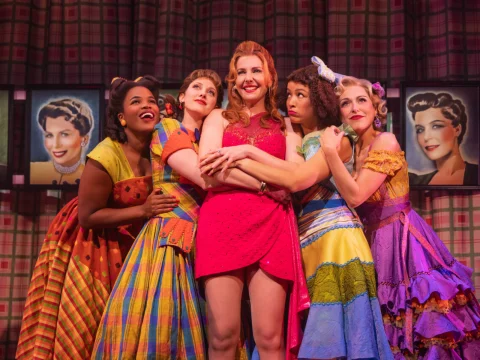 A group of five women in colorful dresses pose together on stage, with framed portraits in the background.