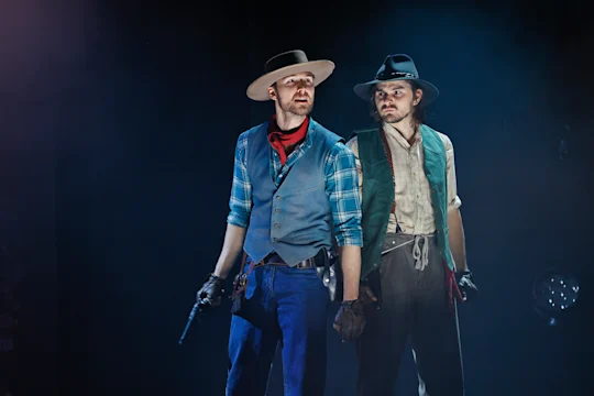 Two men in cowboy attire stand side by side on stage, one holding a revolver, both wearing hats and vests, with dramatic lighting and a smoky background.