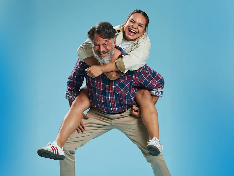 A man gives a piggyback ride to a smiling woman against a plain blue background. Both appear happy and casual.